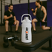 White Per4m water bottle on a gym bench with dumbbells nearby and two men lifting weights in the background