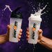 Two clear PER4M shaker bottles in use with frothy liquid splashing out, held firmly in muscular hands against a bold purple MSS-themed background, capturing dynamic energy and movement